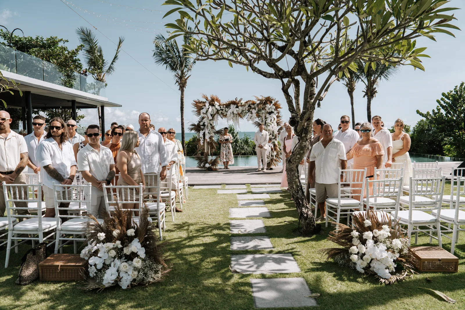 Guests standing for the ceremony at Arnalaya Beach House, frangipani trees over the aisle