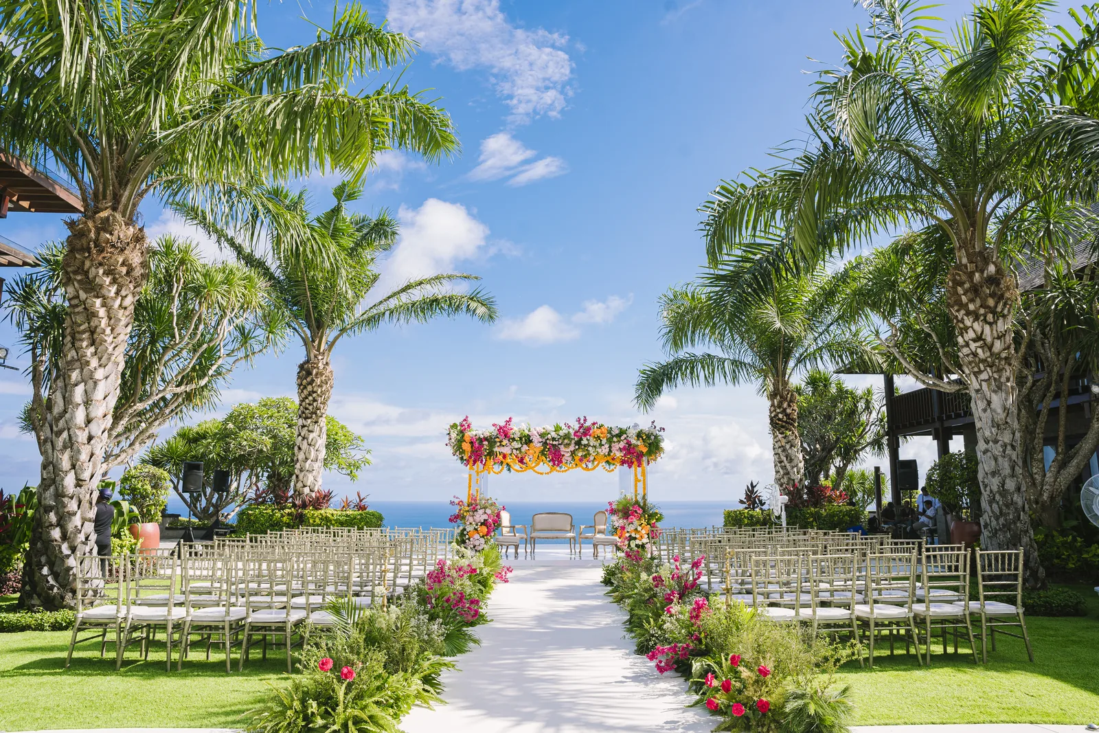 The ceremony aisle at Stones Villas Uluwatu framed by two tall palms in full leaf, the floral arch set against the ocean